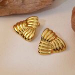 Photo de boucles d'oreilles en éventail doré avec une texture tressée, fabriquées en acier inoxydable, posées sur un fond naturel avec des éléments en bois ou pierre.