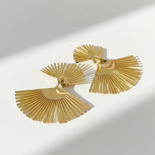Photo de boucles d'oreilles dorées en forme d'éventail, fabriquées en acier inoxydable, suspendues devant un filet d'eau sur fond blanc.
