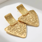 Photo de boucles d'oreilles dorées en forme triangulaire avec une texture martelée, fabriquées en acier inoxydable, sur fond blanc.